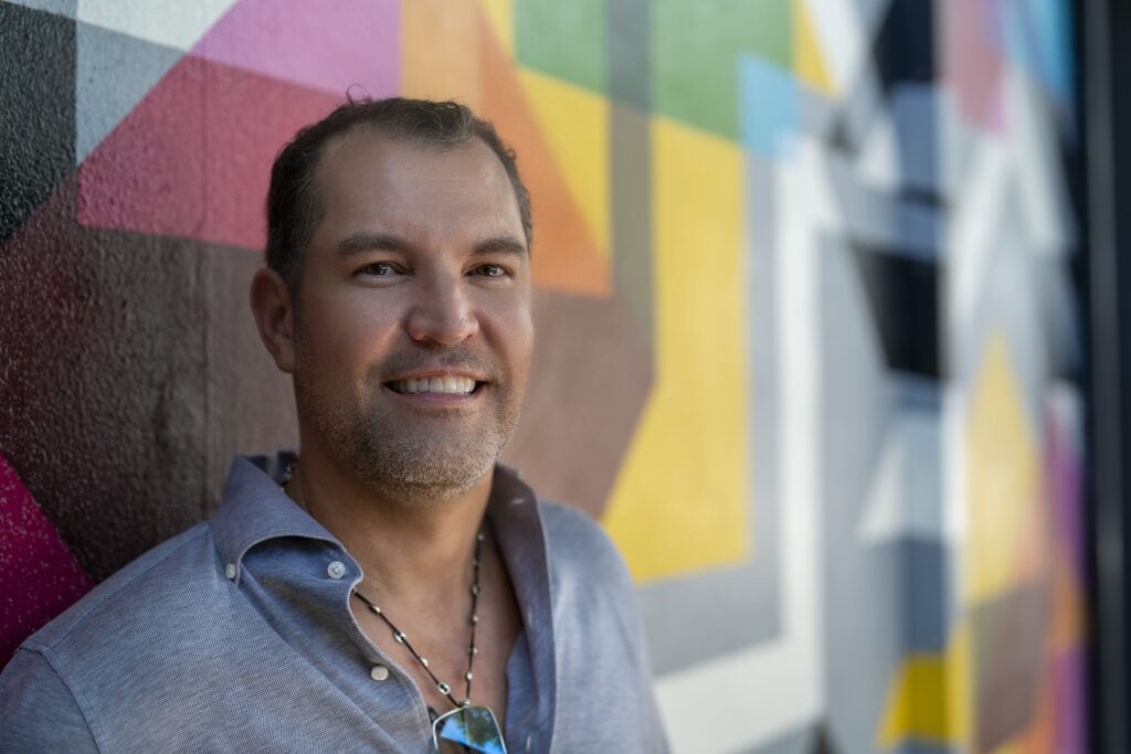 A man smiling in front of a colorful wall.